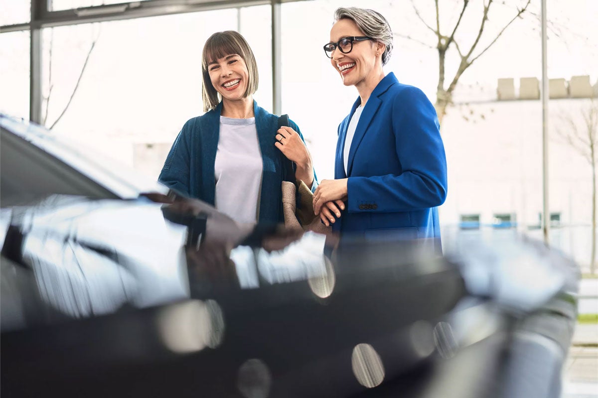 Two women, one with short brown hair and one gray-haired wearing glasses, stand smiling happily in a car dealership.