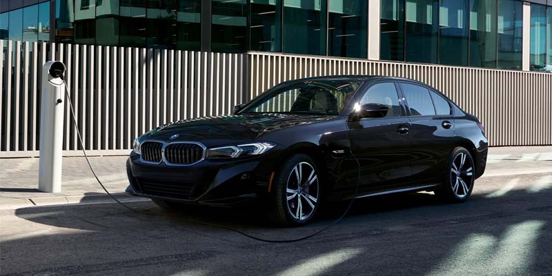 A black BMW sedan is parked beside a public electric vehicle charging station outside a modern glass-fronted building