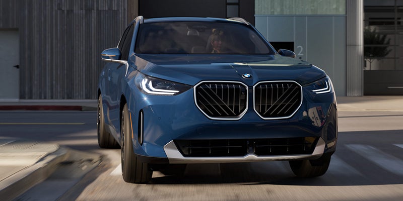 Front view of a blue BMW SUV driving through an urban street, featuring a prominent kidney grille