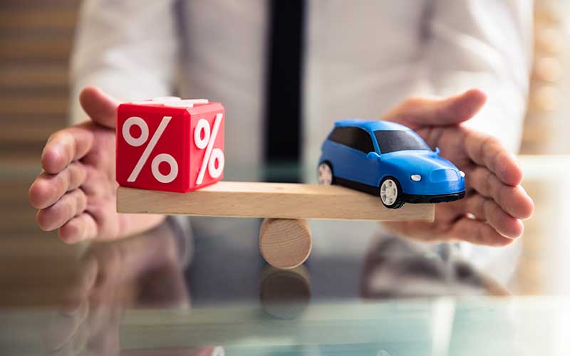 A miniature blue car and a red percentage symbol block are balanced on opposite ends of a wooden seesaw